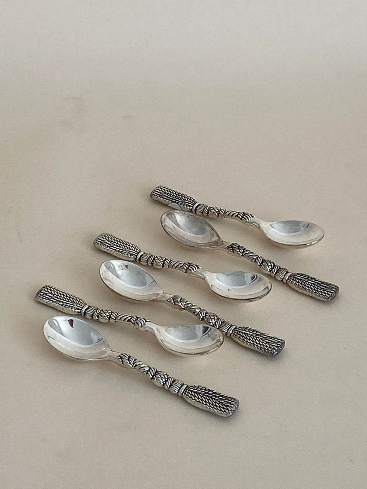 A set of five French Tassel Spoons from Aesthete Label in excellent vintage condition, featuring intricate, textured designs on the handles and arranged in two slightly uneven rows on a plain, light-colored surface. Perfect for a decorative table setting, the polished spoons reflect light beautifully off their surfaces.