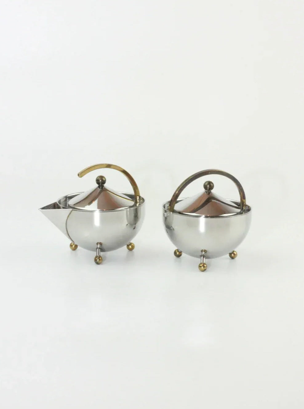 A Danish Sugar and Creamer Set on a white background. One is a stainless steel creamer with a curved handle and spout, while the other appears to be a matching brass sugar bowl. Both have tripod legs with ball-shaped feet and are topped with lids featuring small round knobs. Designed by Danish designer Carsten Jørgensen for Boga Avante Shop.