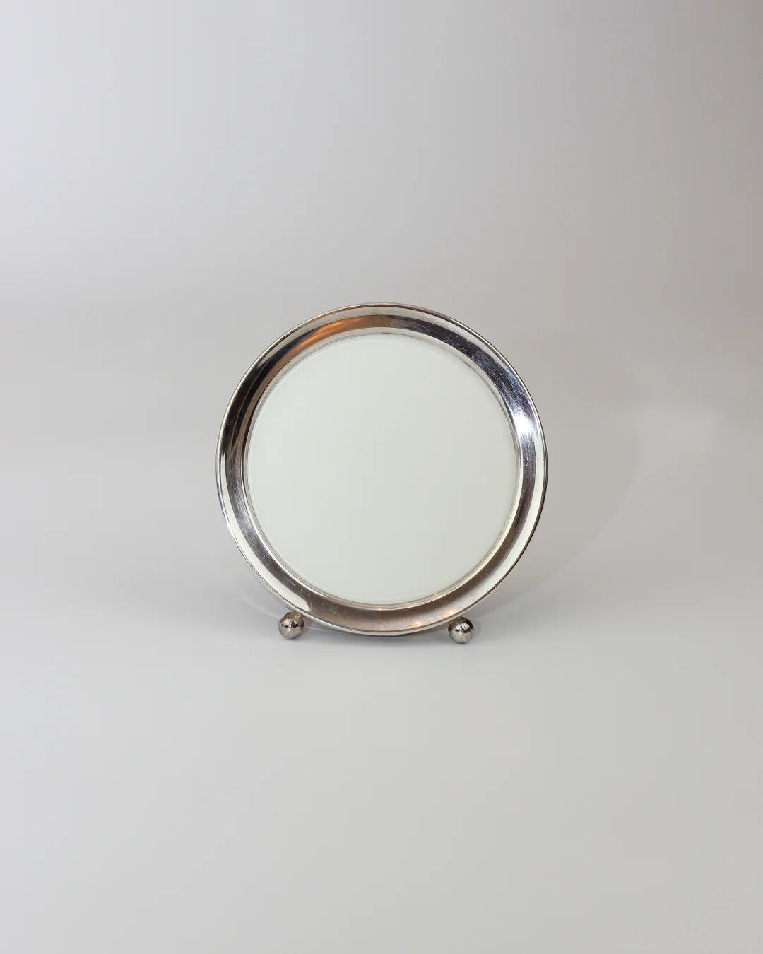 The Round Art Deco Mirror by Boga Avante Shop features a round silver frame and plain white center, standing on two spherical feet against a light gray backdrop.
