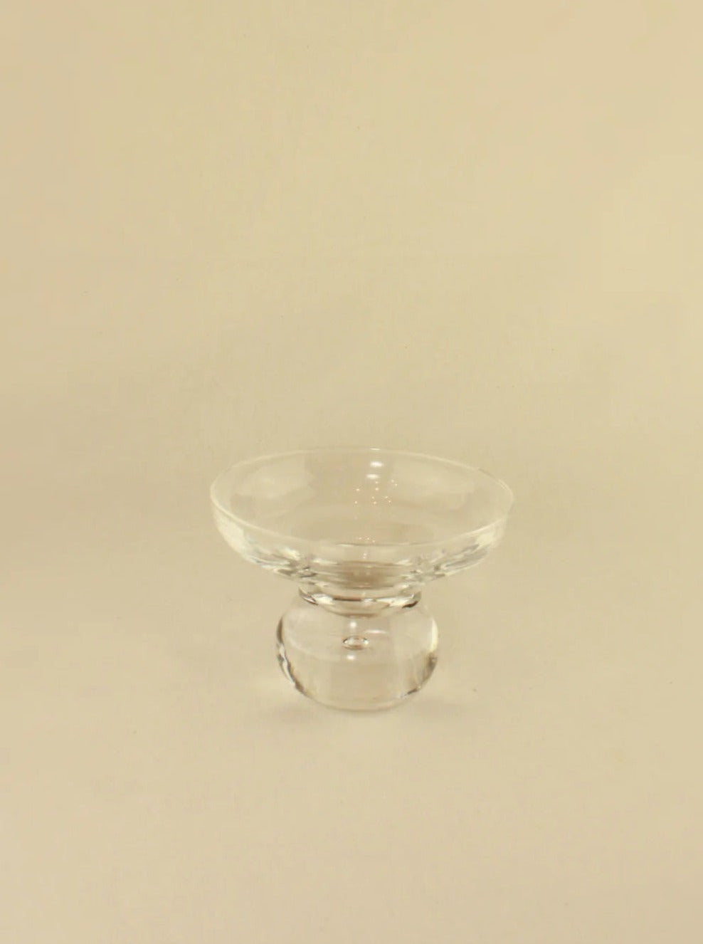 A clear glass pedestal bowl with a round, sturdy base and a shallow, wide bowl on top. Crafted with the elegance of Boga Avante Shop Italian Glasses, the background is a plain, light beige color.