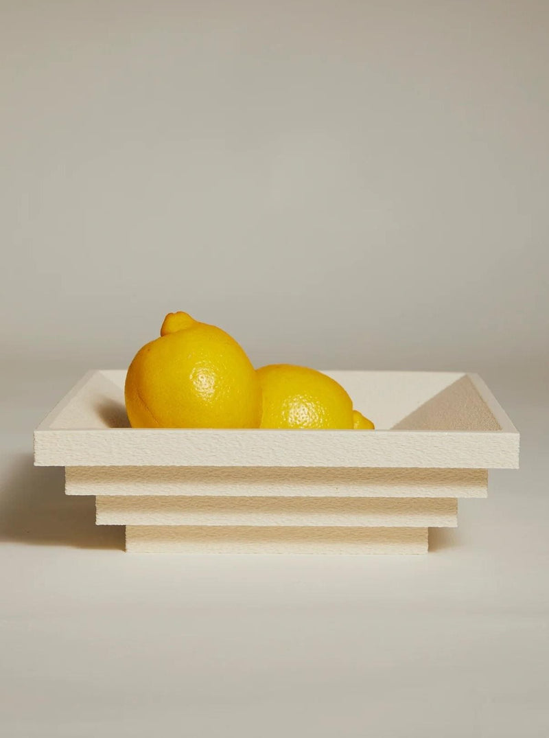 Two lemons sit in the Bau Bowl by Argot Studio, a modern tiered white bowl with a textured surface and layered edges creating a sculptural look. Set against a neutral beige background, the scene highlights the lemons' bright yellow, embodying minimalist elegance.
