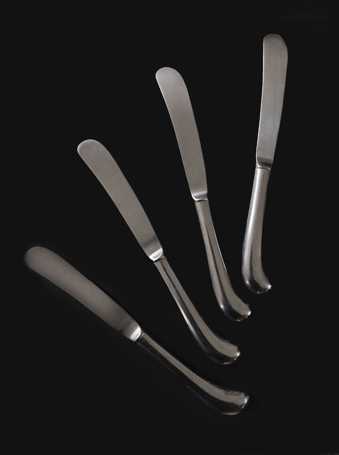 A set of 4 Les Objoies butter knives, crafted from stainless steel with smooth, slightly curved handles, are artistically arranged against a black background, showcasing modern Korean tableware elegance with their rounded ends pointing outward.
