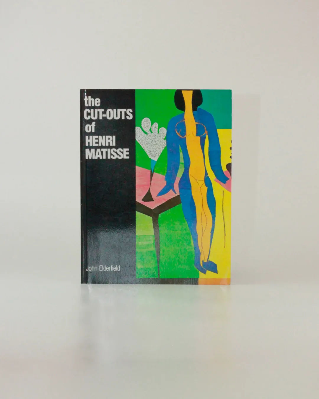 A book titled "The Cut Outs of: Henri Matisse" available at Boga Avante Shop features a colorful cover with abstract artwork. It includes a standing figure painted in blue and yellow and a plant with white leaves on a rectangular surface, set against vibrant green and blue backgrounds, showcasing Matisse's mature cut-outs.