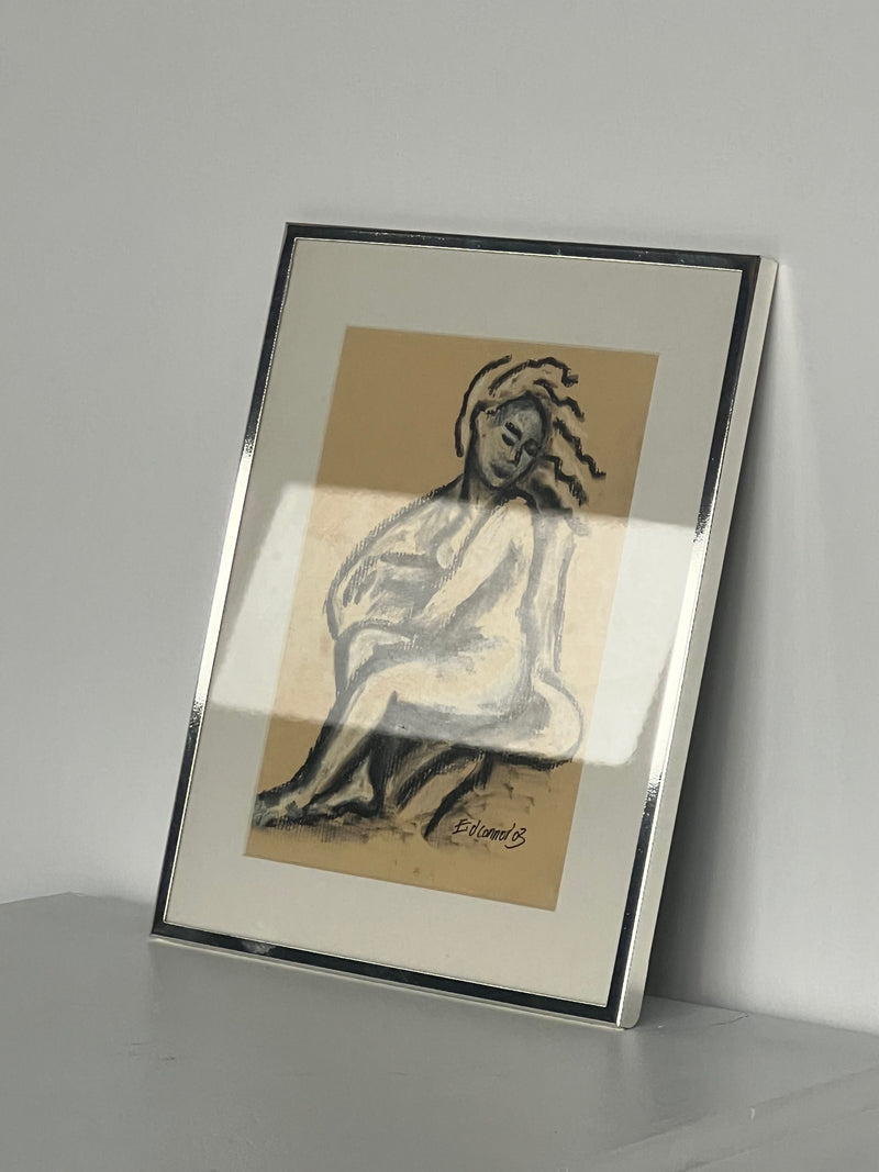 The "Pastel Drawing" by E.D. Connor from Porter Studio features a stylized seated figure, with abstract lines indicating movement. It sits against a neutral beige background, encased in a polished chrome frame and displayed on a smooth surface against a plain wall.