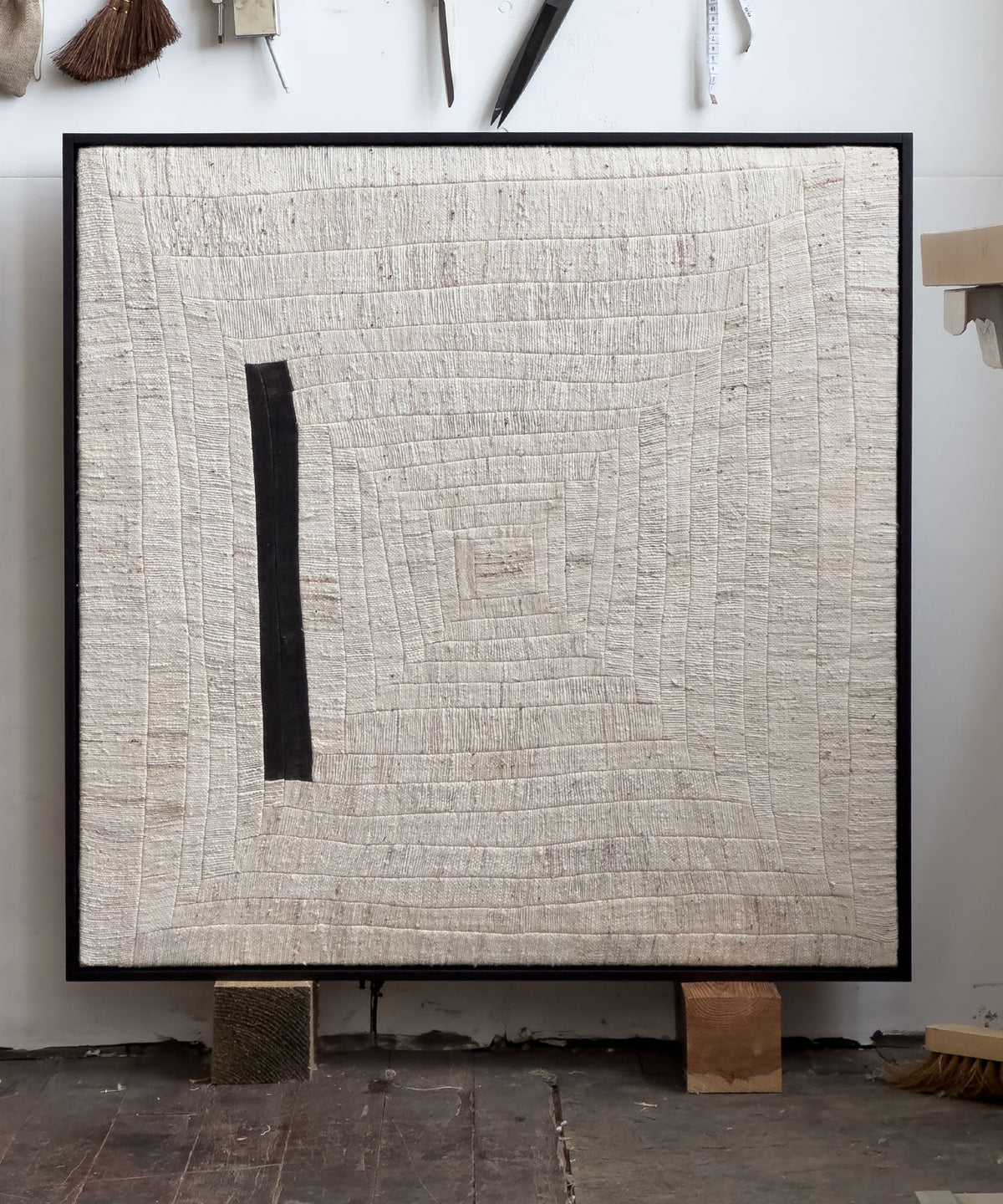 The "to tilt, 2024" artwork by Bec Kirby is a square piece crafted from handwoven kapok fiber cloth. It showcases textured, concentric hand-stitched lines forming a spiral effect on cream-colored fabric, with a black vertical stripe along the left side. This unique artwork is displayed against a white wall and rests on solid oak blocks.