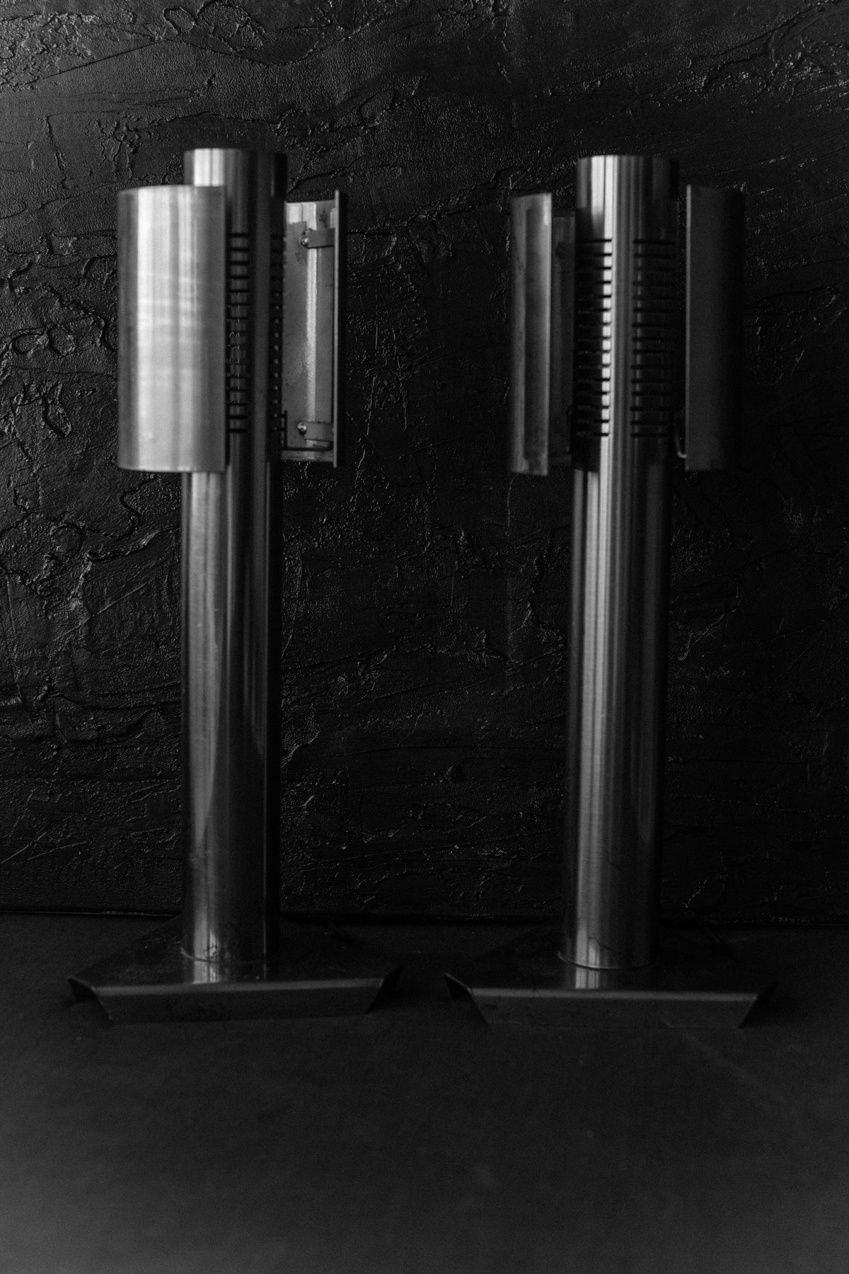 A black and white image showcases Out For Lunch's 1960's Space Age Lamps, featuring tall cylindrical designs with vertical slats. Mounted on square bases, these metallic lamps stand side by side against a textured dark wall, evoking a timeless space-age feel.