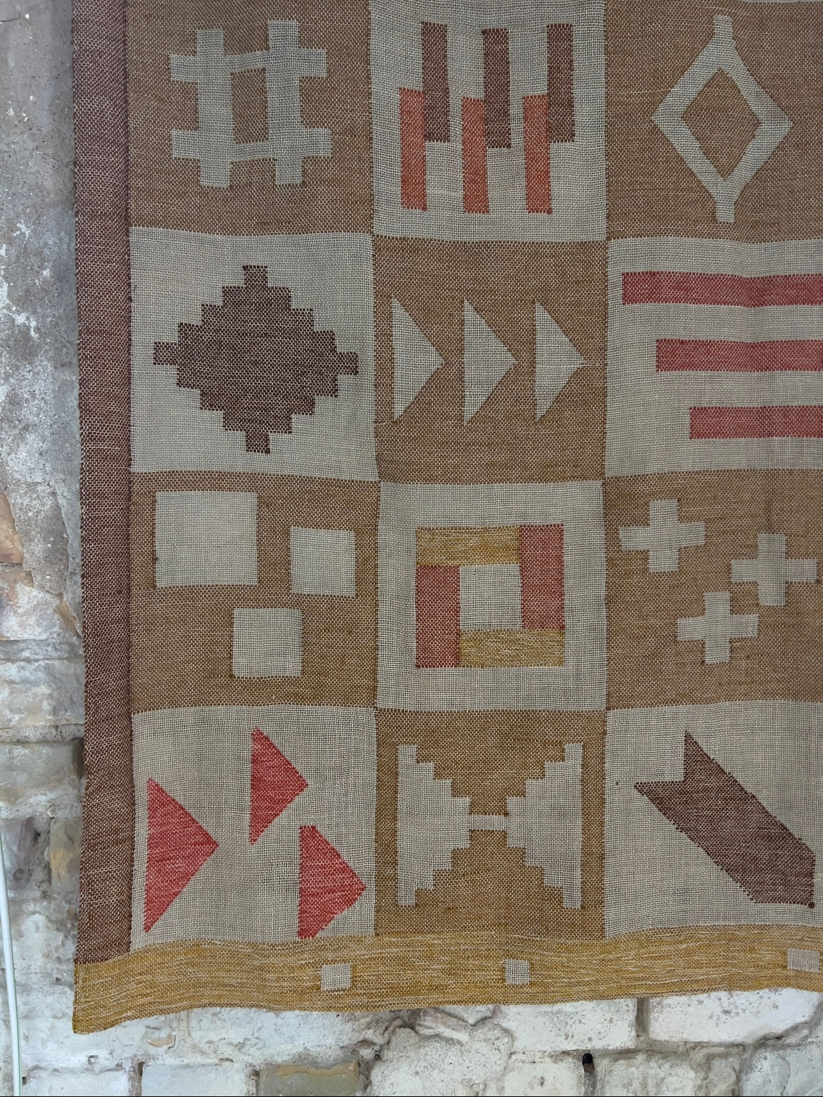 The Large Abstract Wool Wall Tapestry by Veter Vintage (Sweden, 1988) features a handwoven grid of unique geometric squares in earthy brown, red, and tan tones, beautifully displayed against a rustic stone wall.