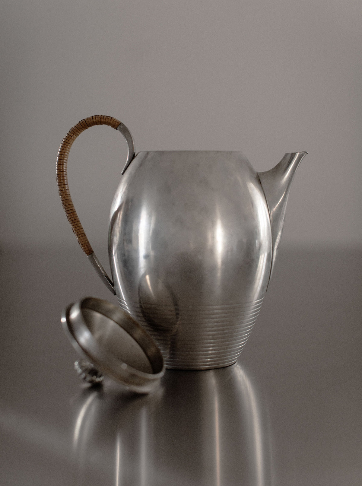 The Out For Lunch Art Deco Teapot, featuring a vintage pewter finish, a curved spout, and a round body, is elegantly displayed on a reflective surface. Its handle is wrapped in brown material, blending style with functionality. The lid is placed next to the teapot, enhancing the dining experience against a neutral background.
