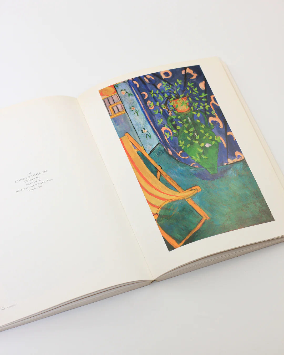 The Henri Matisse Book from Boga Avante Shop lies open on a white surface, showcasing a vibrant painting of a yellow chair beside a blue, leafy curtain, echoing Matisse's modern art style. The right page highlights this Fauvism-inspired piece, while the left page contains text.