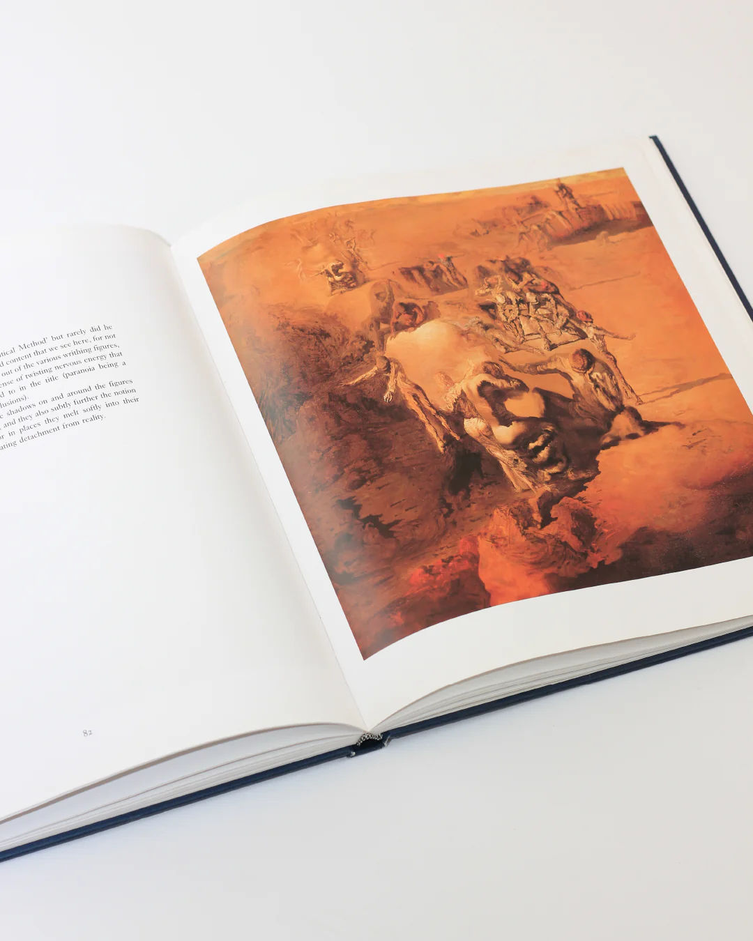 On a white surface lies the "Dali The Masterworks Book" by Boga Avante Shop, open to reveal a surreal, colorful painting with abstract forms in earthy tones reminiscent of Salvador Dalí. Text fills the left page, while the right page highlights this vibrant modern art piece.