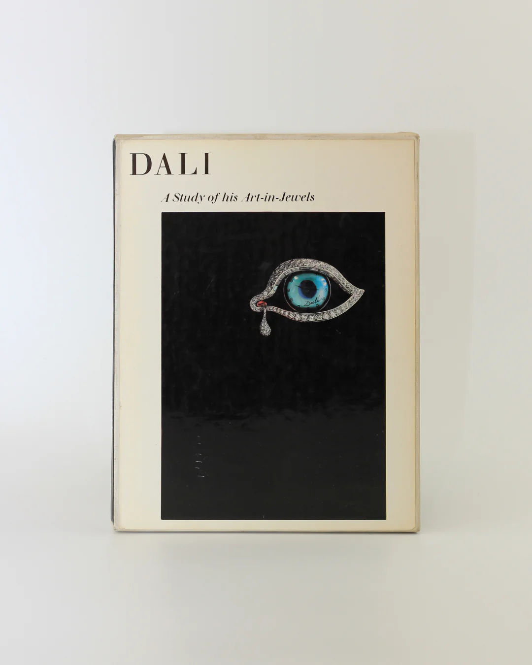 The book cover, "Dalí: A Study of his Art-in-Jewels" by Boga Avante Shop, showcases a surrealist jewelry image featuring an eye-shaped jewel with a blue iris and teardrop. The contrasting black background enhances Dalí's artistic legacy through this captivating design.