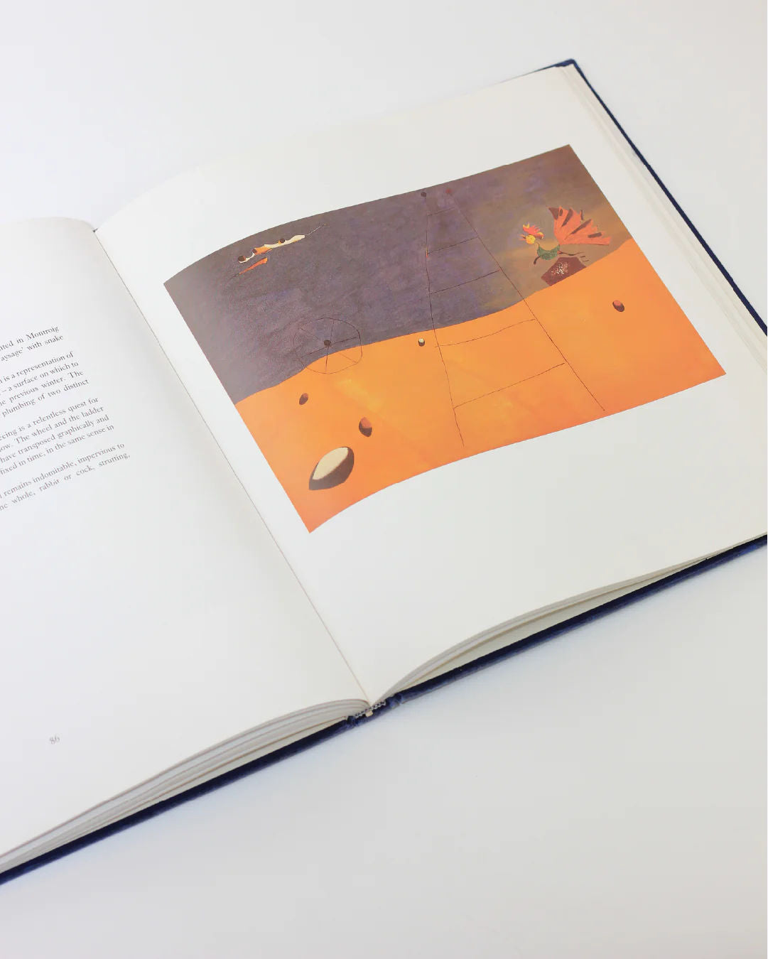 The Mirò The Masterworks Book by Boga Avante Shop showcases an open page with a minimalist landscape in orange and purple geometric shapes on the right, inspired by Joan Miró's surrealism. The left page features text embodying modern art's essence.