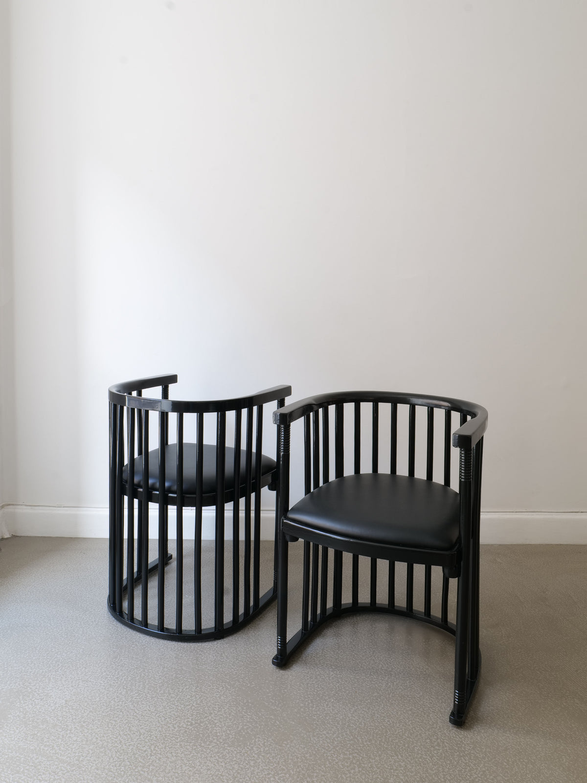 Two Bentwood Armchairs from Collection apart, featuring lacquered black bentwood frames and slatted backs, sit on a light gray floor against a plain white wall. These chairs have cushioned seats with black leather upholstery and showcase unique open designs reminiscent of the Josef Hoffman style.