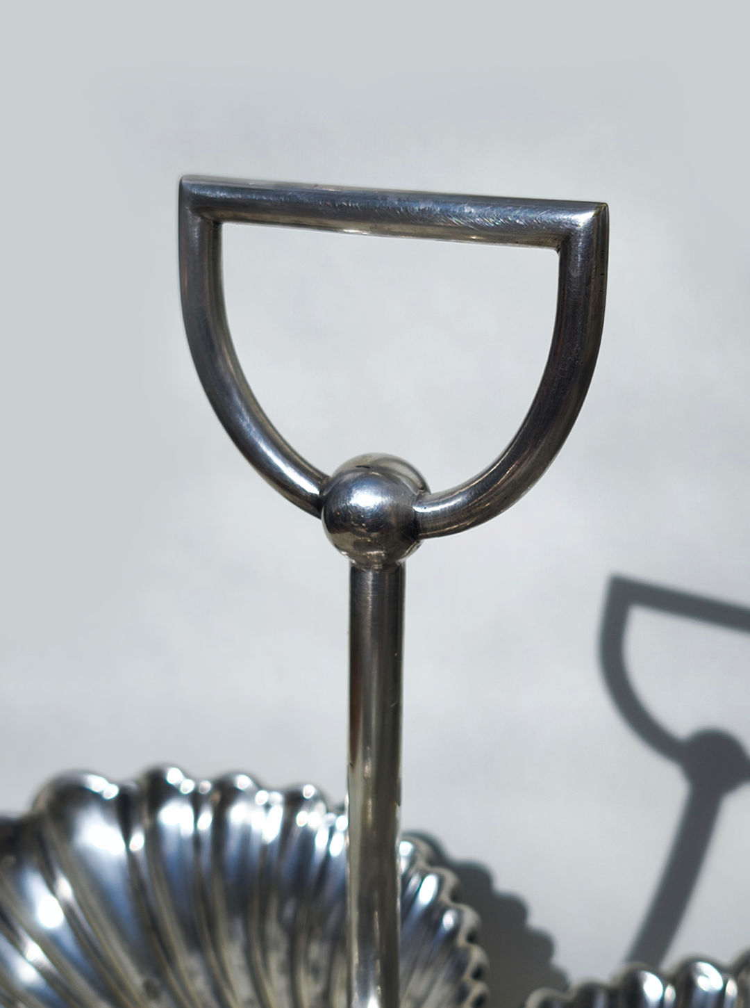 A close-up shows the polished silver handle with a semicircular design on the Les Objoies Large Silver-plated Triple Tray, which features shell-shaped compartments. The handle’s shadow appears on a light gray background.
