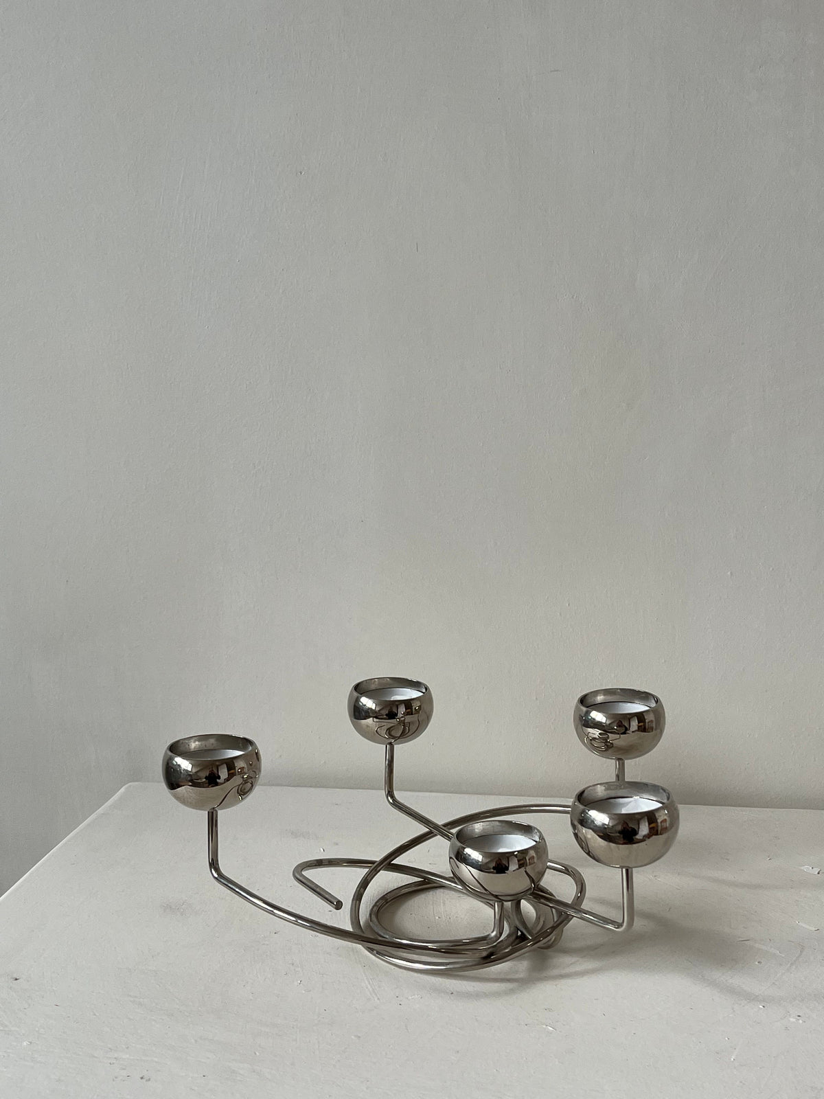 Introducing the Chrome Candleholder by septembre studios: a modern, sculptural piece featuring five rounded holders on an elegantly intertwined base. Its unique angles stand out beautifully against a plain gray background on a white surface.