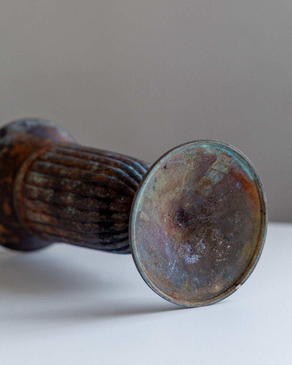 A close-up of the Spigel Vintage Cast Iron Vase reveals a ribbed metal design with a round, flat end, resting on a white surface. The vase displays an antique charm with blue and brown corrosion patterns, evoking the essence of its vintage character against the blurred gray background.