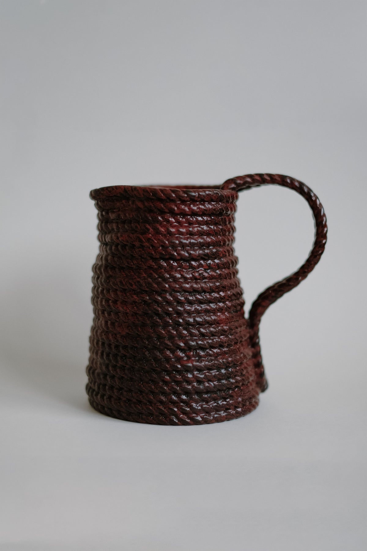 The Handmade Burgundy Vase by septembre studios features a thick, coiled rope design with a matching textured handle, crafted in ceramic with a rich burgundy finish and shown against a plain light gray background.