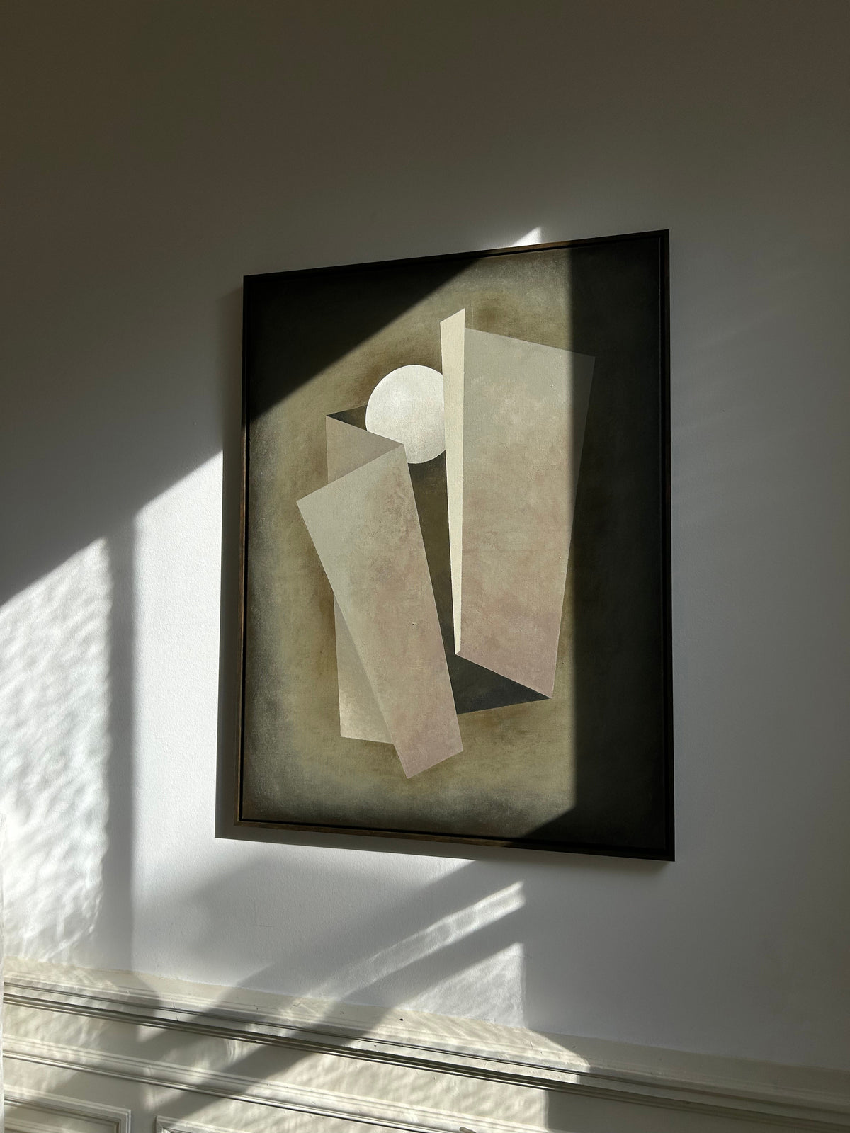The 'Unfolding' Painting by Iris Bavelaar, an abstract acrylic on linen with geometric forms and a white circle, is displayed on a wall where angled sunlight casts sharp shadows across the artwork and the surrounding space.