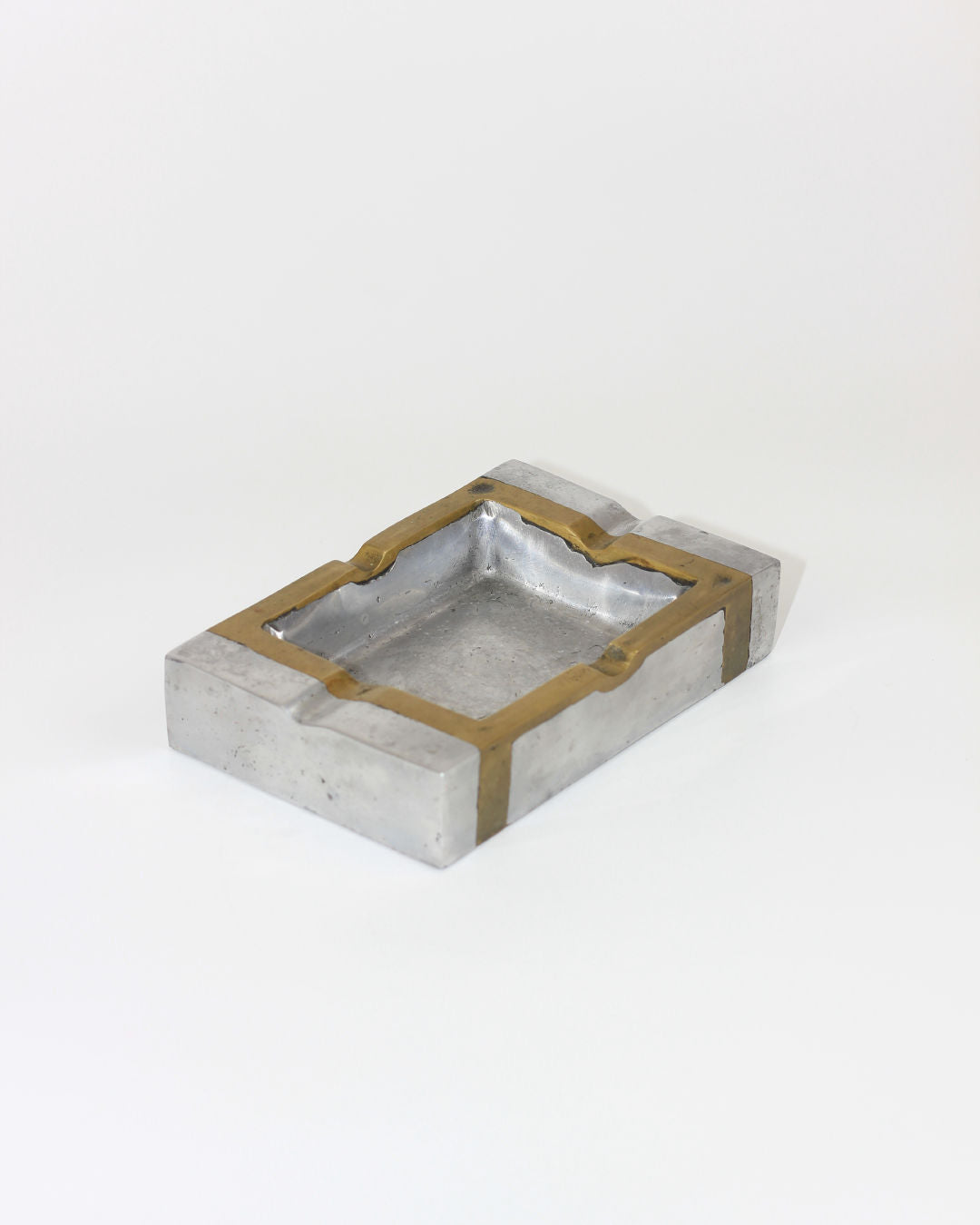 The Brutalist Ashtray 70s by Boga Avante Shop has a rectangular silver body with gold trim and side grooves for cigarettes, made in Spain. Seen on a plain white background.