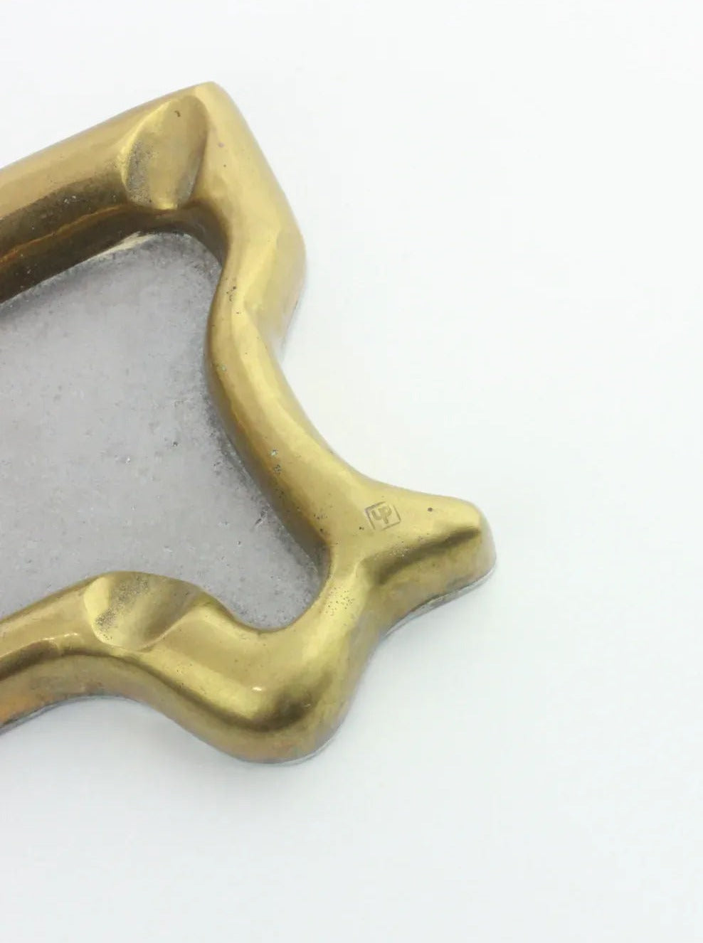 A close-up view of David Marshall's brass and aluminum object with an abstract shape and smooth, rounded edges. The surface shows a polished finish with some wear. A small engraved mark or logo is visible on the right side of this Brutalist Ashtray by Boga Avante Shop. The background is plain white.