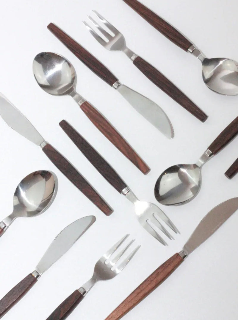 An assortment of silverware, including stainless steel knives, spoons, and forks with dark teak handles, is arranged in a diagonal pattern on a white surface. The Boga Avante Shop Mid-Century Cutlery Set features polished metal and sleek design.