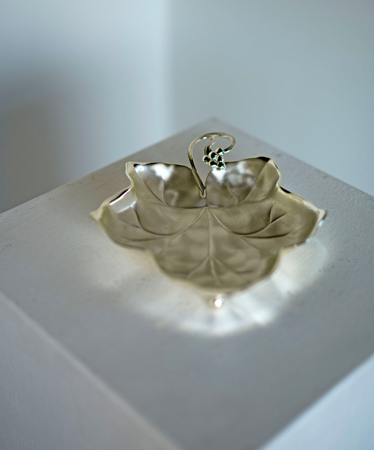 A Grape Leaf Dish by Porter Studio, showcasing intricate vein details reminiscent of the 1930s, is elegantly displayed on a white square pedestal against a plain background. Its soft shadows and timeless design evoke an era of refined taste.