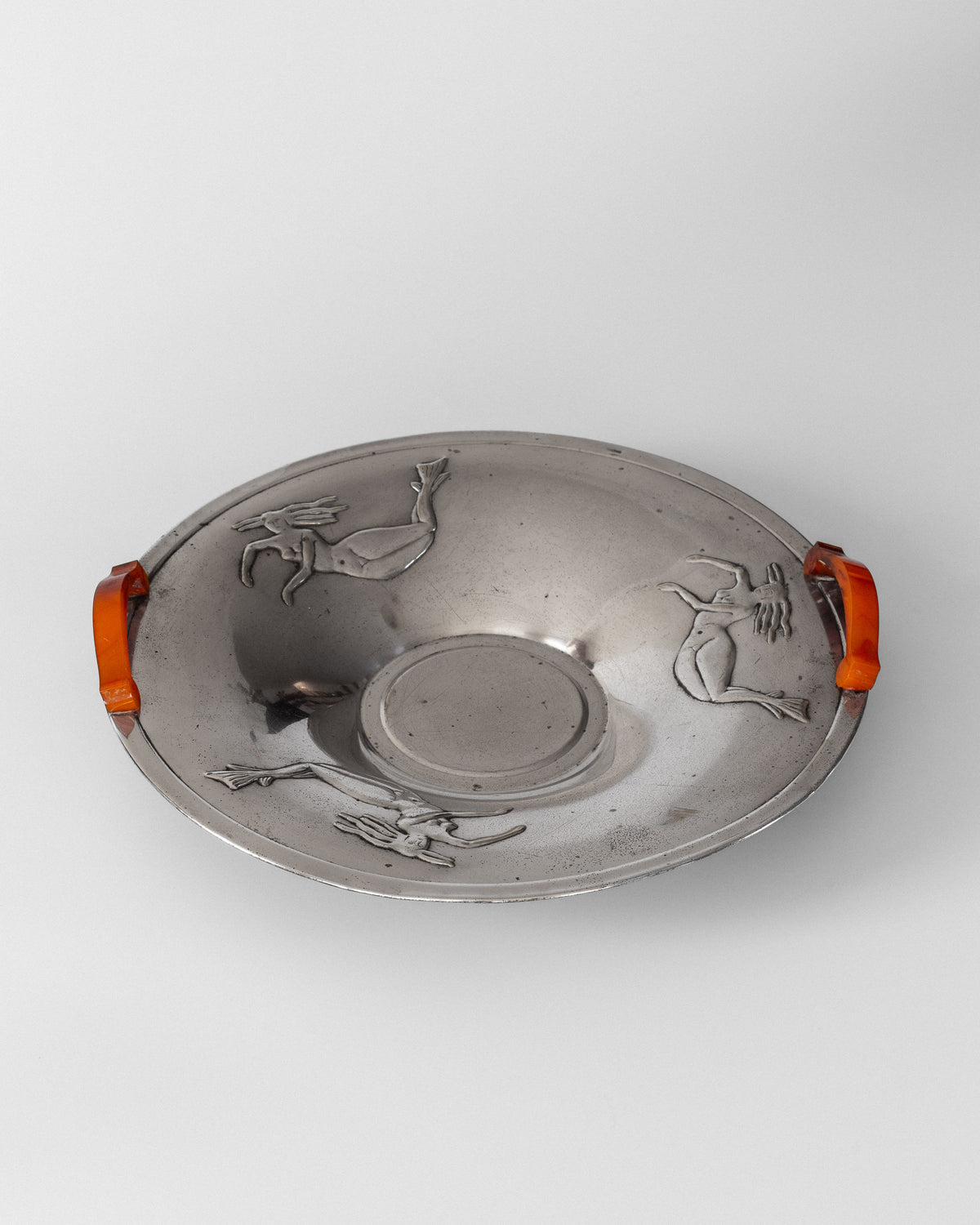 The Decorative Bowl with Mermaids from the 1930s by Spigel is a round, silver-plated piece featuring two orange handles and engraved mermaid designs. It has a central circular depression and exudes a vintage, artisan charm reminiscent of the Swedish Grace style, all set against a plain white background.