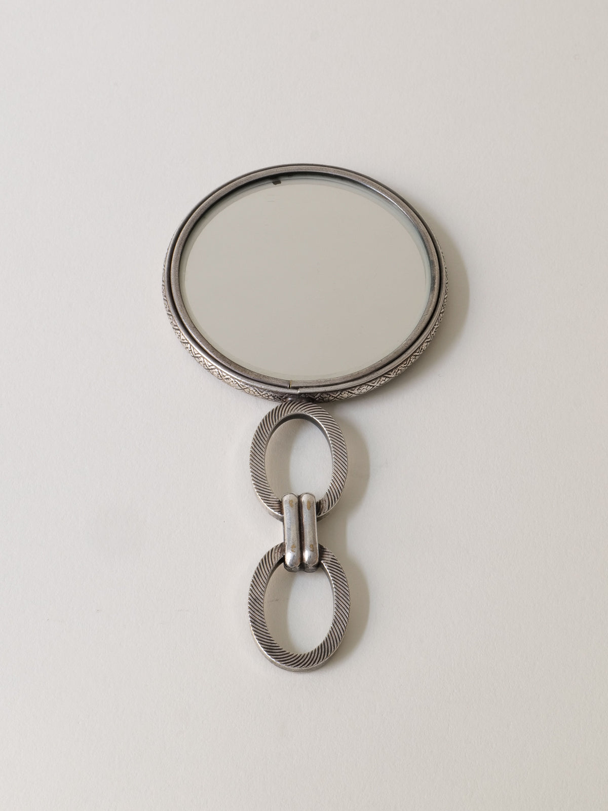 The "Hand Mirror Jean Després 1920s" by Collection apart features a round mirror with a decorative metallic handle, showcasing two interlocking rings with a textured design, evoking Art Deco flair inspired by Jean Després.