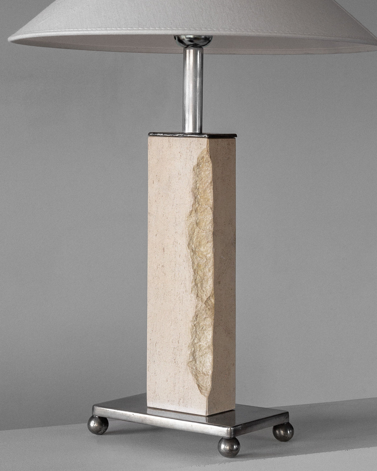 The Spigel Modernist Travertine and Metal Table Lamp features a rectangular stone base with a vertical groove, an Art Deco metallic stand with four round feet, and is topped by a light grey conical shade, adding elegance to any room.