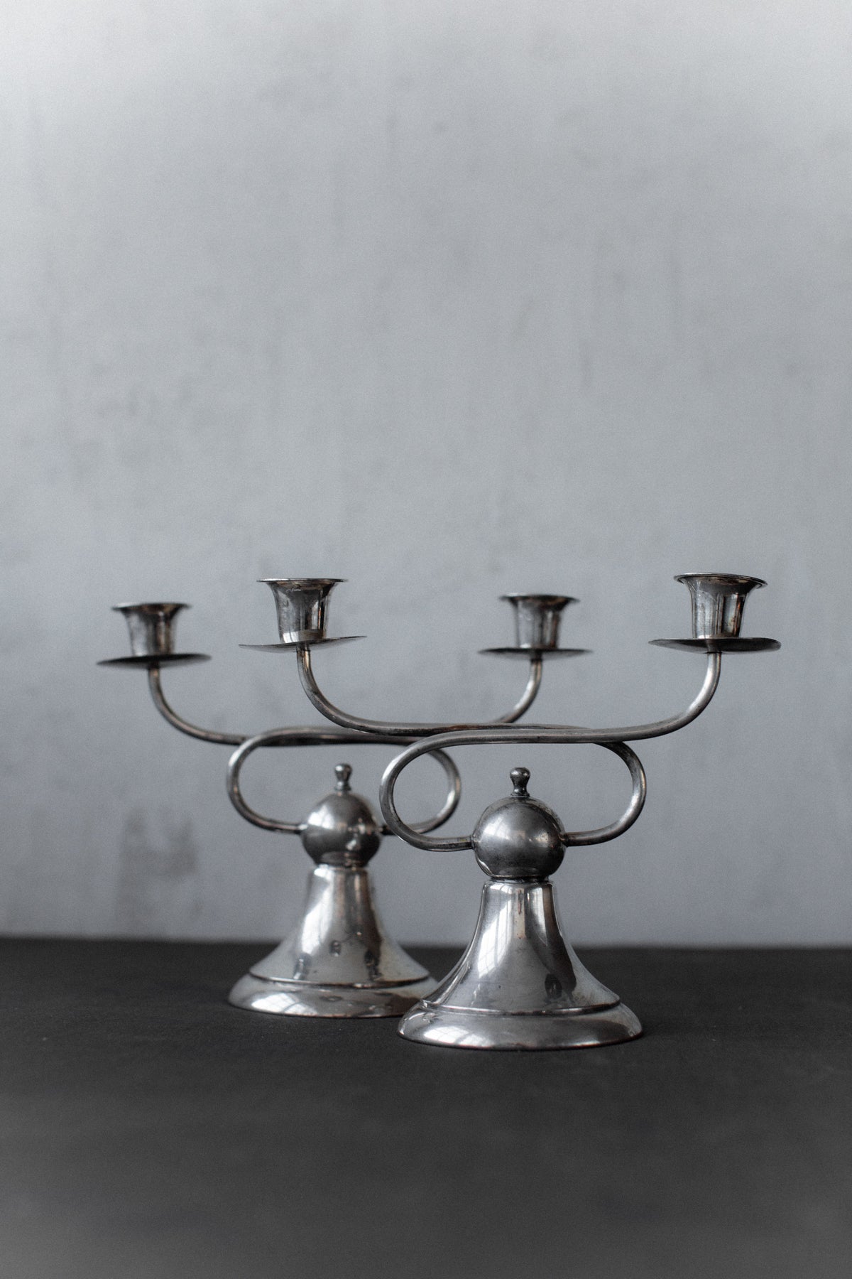 Two Art Deco Candleholders by Out For Lunch, each with three arms, sit on a dark surface against a light gray background. Their geometric elegance and minimalist lines reflect vintage Art Deco style.