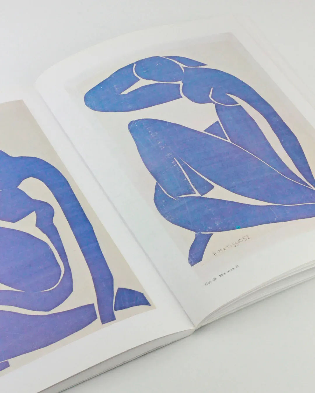 An open book titled "The Cut Outs of: Henri Matisse" from the Boga Avante Shop showcases abstract blue papercutting art forms of human figures. The left page reveals a partial figure with an extended arm, and the right page presents a crouching figure with a hand on its head. The minimalist design, evocative of Matisse's mature cut-outs, highlights fluid lines and shapes.