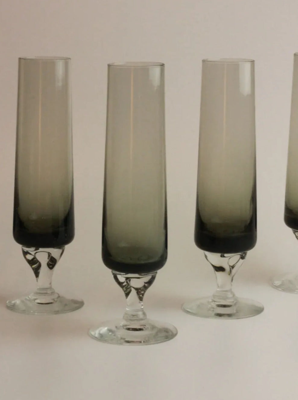 Four tall, slender, smoky grey champagne flutes with vintage flute-shaped design are arranged side by side on a neutral background. The flutes have a gradient design, becoming darker towards the base. This is the Set of Five Vintage Flute by Boga Avante Shop.