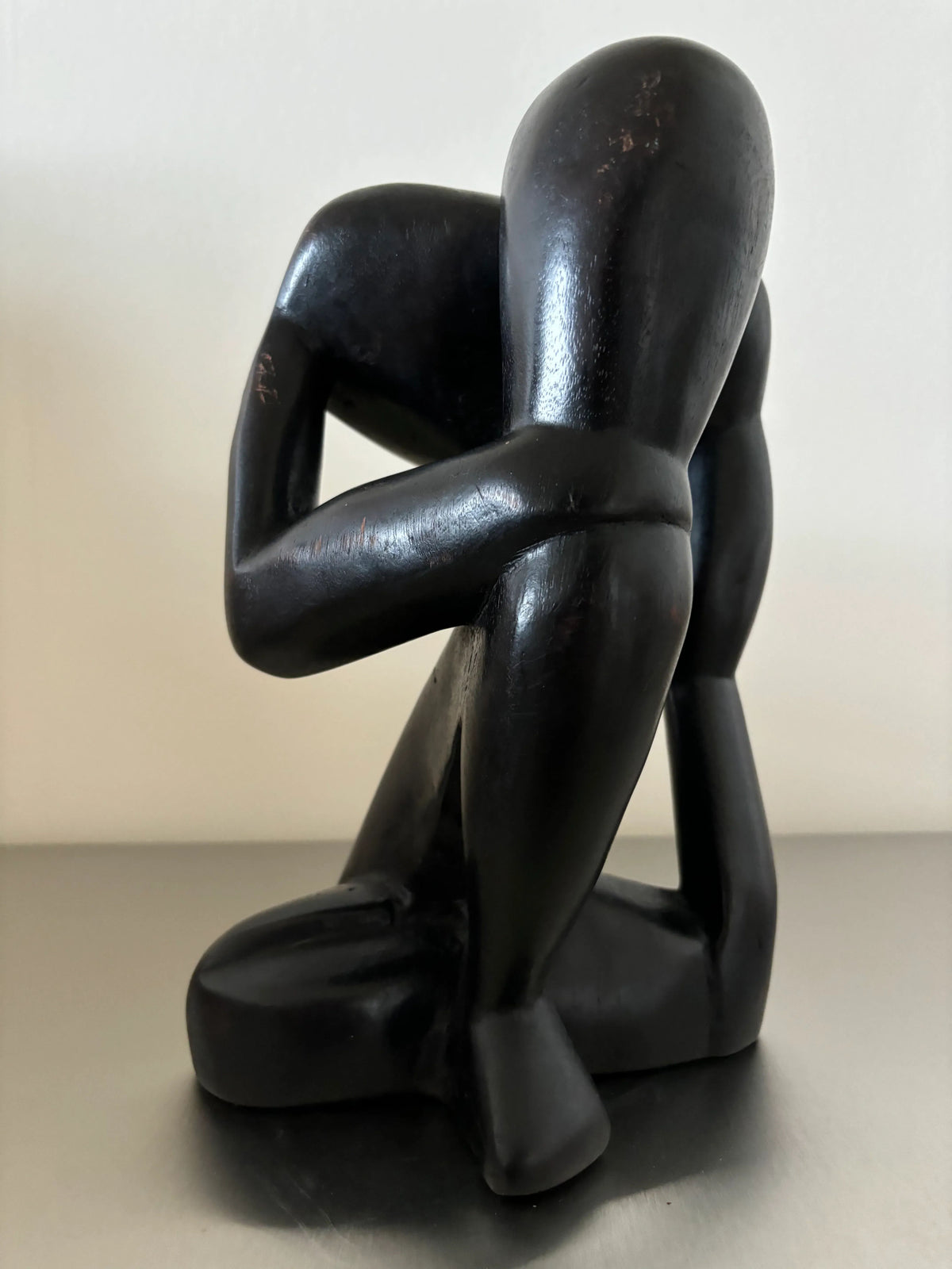 A CU-RA-TED Black Wooden Object - “The Thinker,” a sculpture of a human figure kneeling with its head resting on its knee, arms wrapped around its legs. The craftsmanship highlights smooth, flowing curves and simplified human forms, enhanced by a sleek black finish.