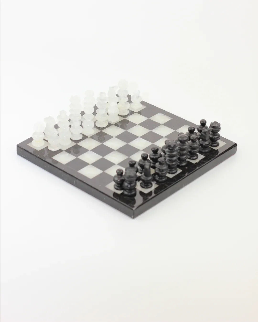 The Handmade Chess Board in onyx by Boga Avante Shop features an elegant setup with hand-carved black and white glass pieces. The onyx stone board contrasts beautifully with the translucent white and solid black figures, viewed artistically from above.
