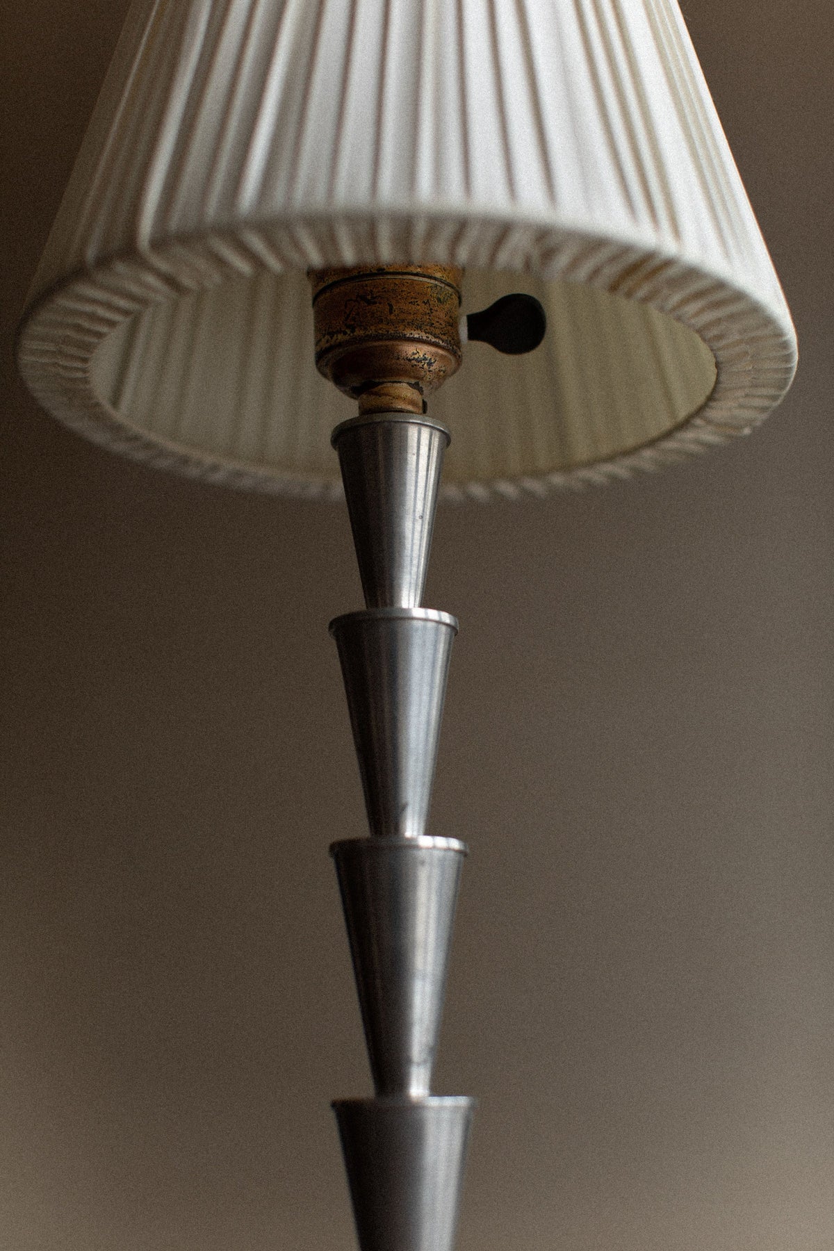 A close-up of an Out For Lunch 1920's Swedish Grace Lamp with a pleated white lampshade. The lamp's base features a stack of tapered, metallic cones, giving the vintage product a sleek and contemporary look against a neutral background.