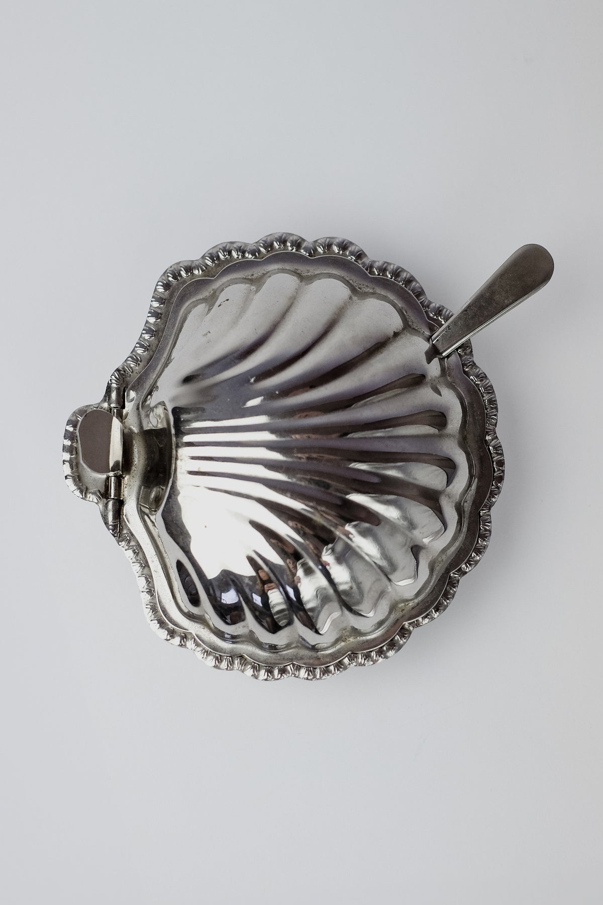 Elevate your dining with the Shell Shaped Butter Dish with Spoon by septembre studios. This seashell-inspired silver dish, viewed from above, features a scalloped edge and shiny, reflective surface.