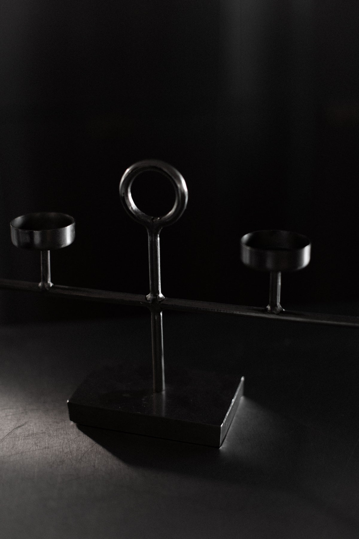 A dark, minimalist photo features the "Steel Candleholder" by Out For Lunch, showcasing two circular holders flanking a central upright stem. This geometric design on a flat surface creates dramatic shadows and light.