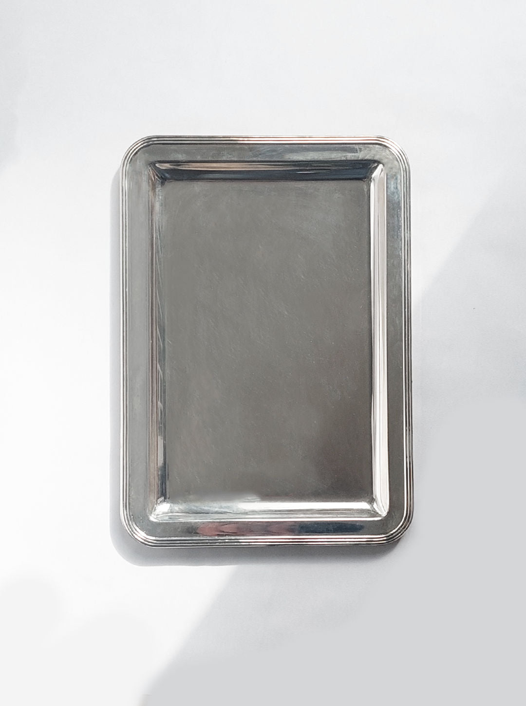 The Les Objoies Silver-plated Rectangle Tray, featuring a raised edge, is showcased against a plain, light background. This minimalist tray boasts a clean, shiny surface and a simple, classic design. Centered in the image, it casts a subtle shadow to one side due to the lighting.