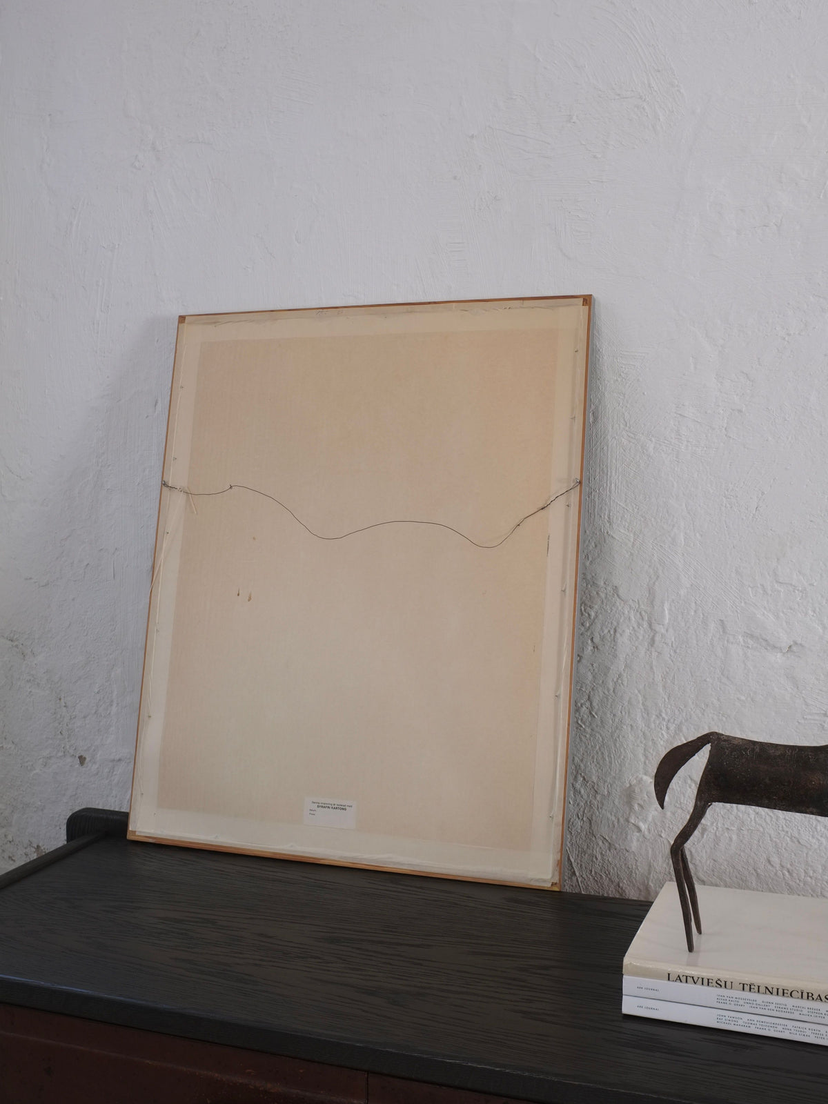 The framed Ebba Von Wachenfeldt Charcoal Drawing 1883 from Veter Vintage leans backward against a plain white wall. Below, a textured dark wooden surface displays a book and a claw-like sculpture. The minimalist setting is dimly lit.