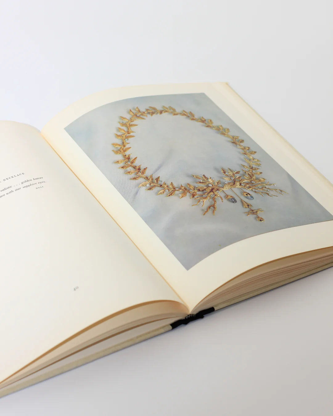 The book "Dalí: A Study of his Art-in-Jewels" by Boga Avante Shop is slightly ajar on a white surface, showcasing a photograph of an ornate gold necklace reminiscent of Dalí's surrealist jewelry across its pages.