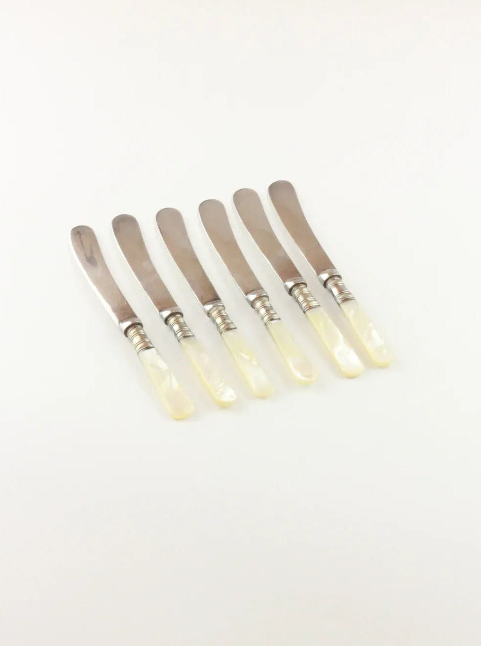 A set of six Mother of Pearl Knives from Boga Avante Shop arranged in a row on a white background. Each knife boasts a shiny, metallic blade and a pearlescent mother-of-pearl handle, reminiscent of vintage flatware. The knives appear elegant and uniform in design.