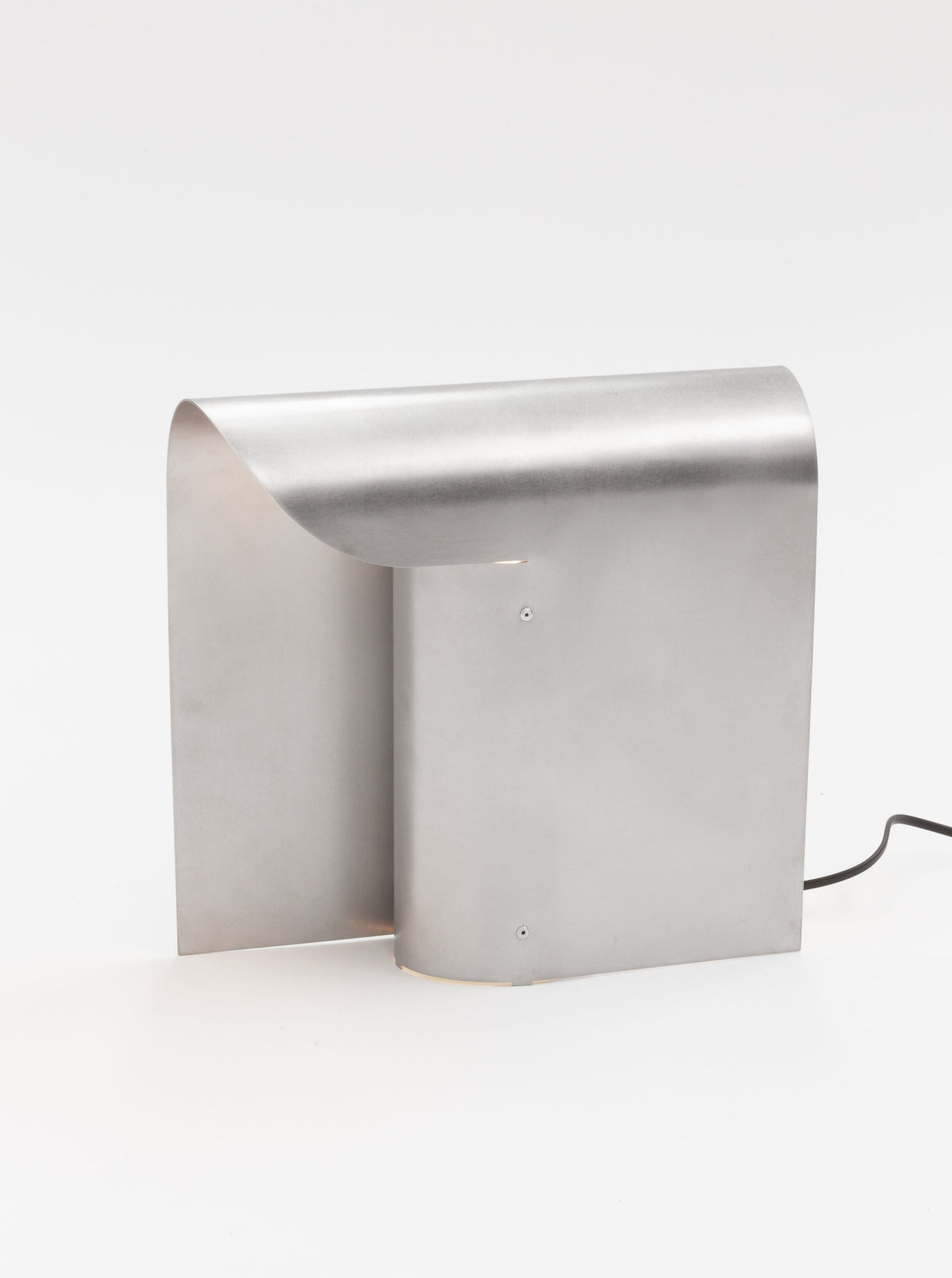 A sleek, modern metallic light fixture with a minimalist design, featuring a curved sheet of brushed stainless steel standing upright. The elegant and contemporary appearance is complemented by an E27 socket for versatile bulb options, with a black electrical cord partially visible on the right side. This is the Arc Table Lamp by Paul Coenen.