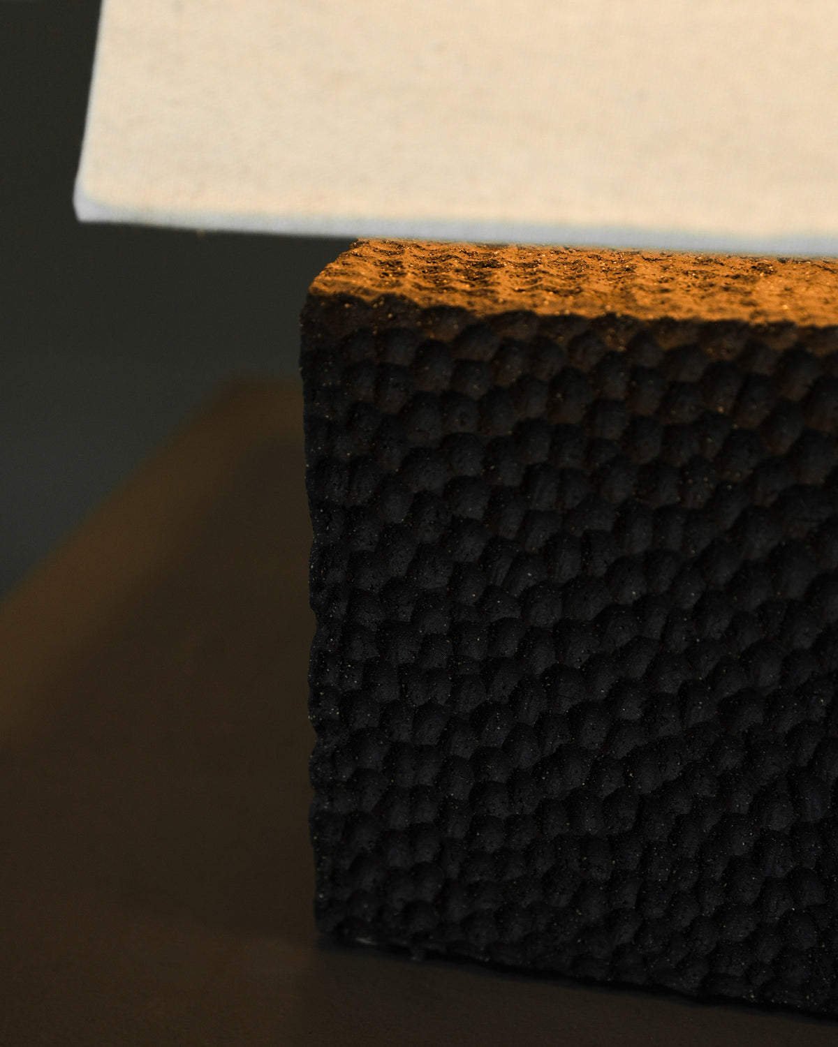 Close-up of Studio OSKLO's Akune Ceramic Lamp - Number Five, a black, textured rectangular base with a dimpled surface, supporting a sleek white element on top, shown against a dark background.