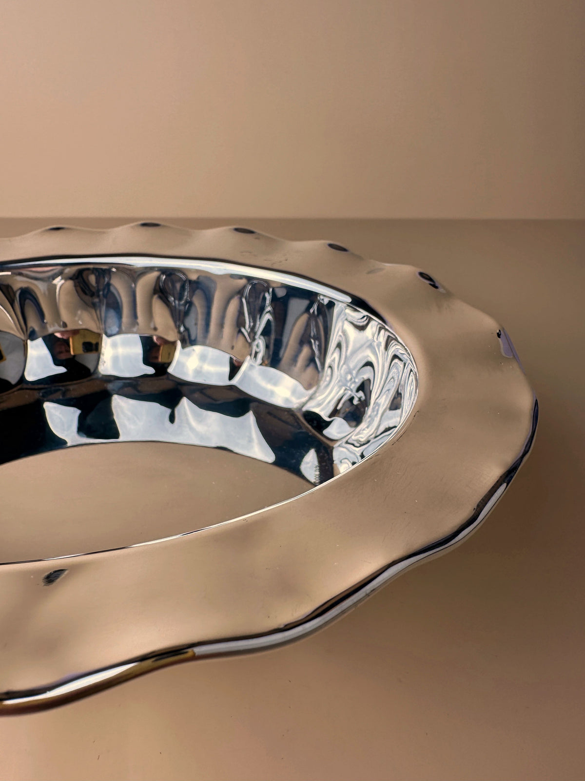A wavy-edged, silver-plated Italian footed bowl from Dodo Vintage is placed on a flat, beige surface. The reflective surface of this Mid Century silver plated Italian bowl captures ambient light and shadows, adding depth and texture to the image. The background is plain and unobtrusive, highlighting the bowl's elegant design.