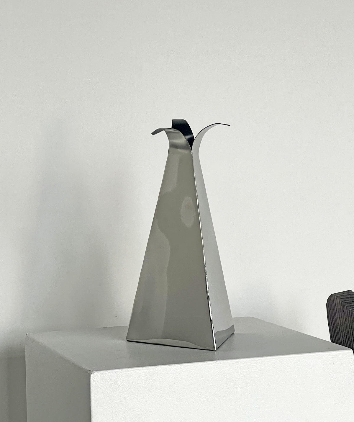 A sleek and modern Triangular Vase by Porter Studio with an angular design rests on a white pedestal against a plain wall. Its reflective surface captures subtle shadows and contours, adding an artistic touch to the minimalist setting, making it perfect for holding flowers or serving as a stylish decorative piece.
