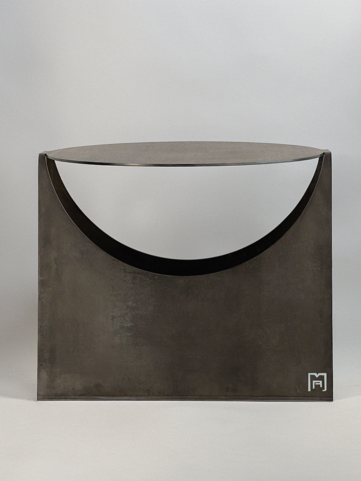 The 'Maru' Side Table by Modan features a minimalist, geometric steel design with a crescent-shaped top, shown on a plain background. Its smooth finish and modern industrial style are accented by a small white Modan logo in the corner.