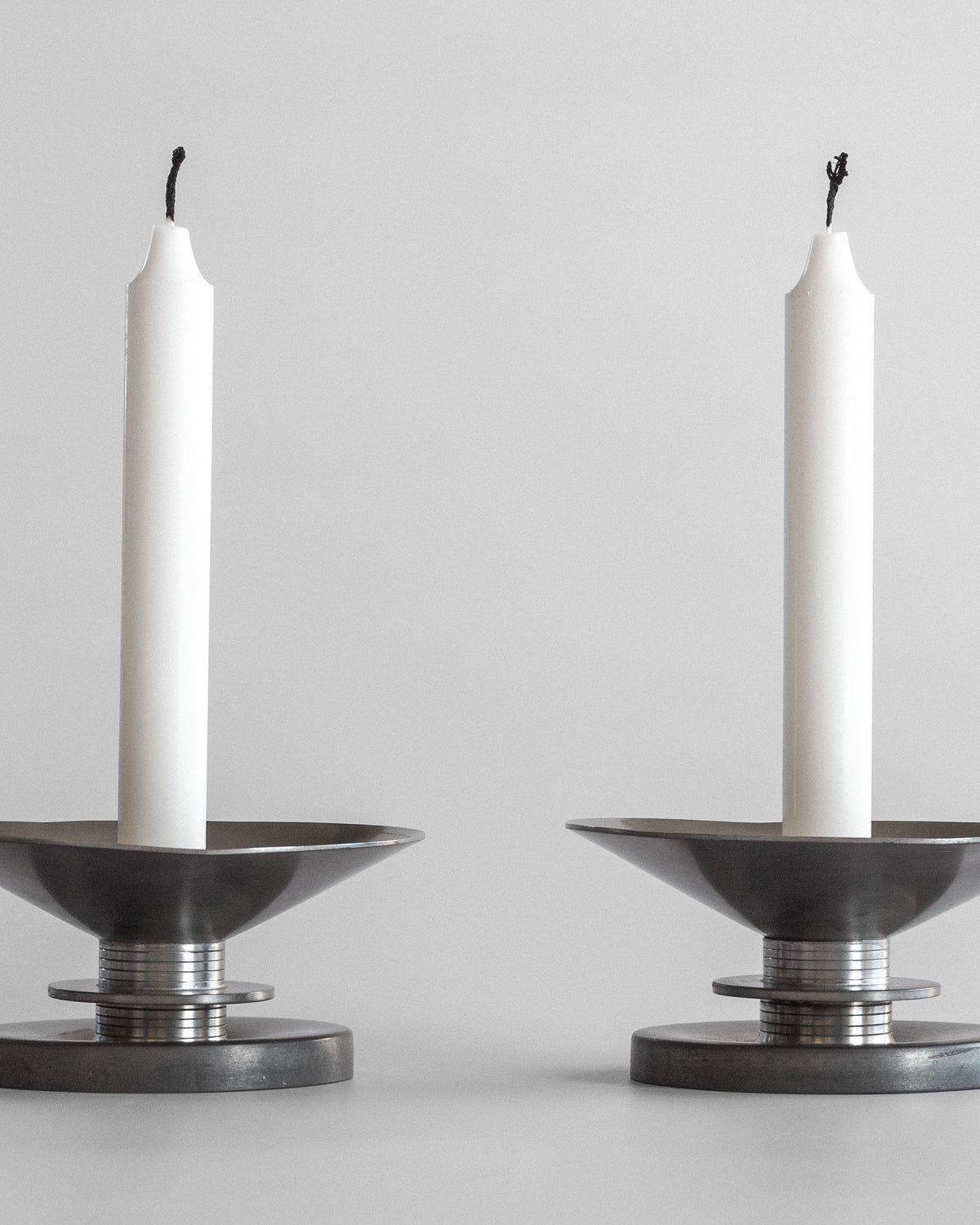 Two unlit, white candles in Spigel's Sylvia Stave's 1930s Modernist Candleholders stand against a plain, light gray background, capturing the essence of 20th-century decorative arts with their elegant pewter design and wide, circular bases.