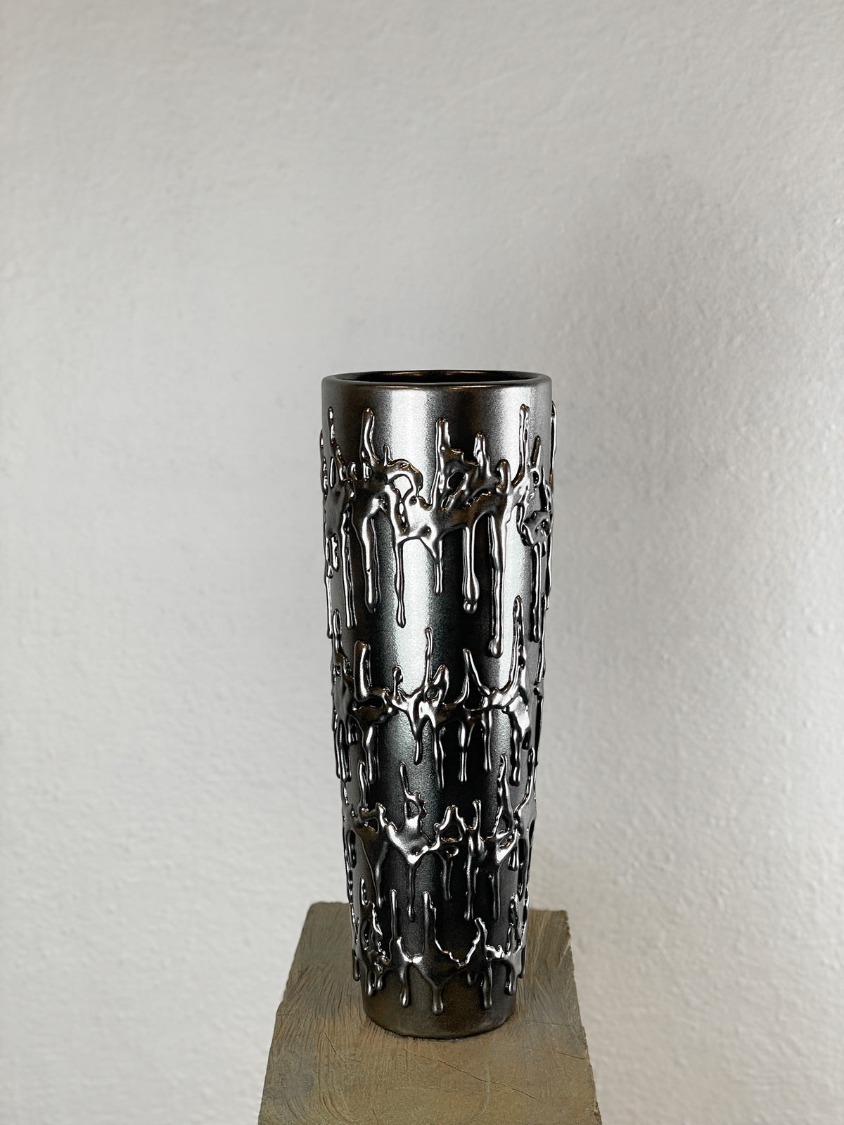 The Almost Chrome Vase .001 by John Nordenstein is a tall, chrome vase with a shiny finish and abstract raised dripping patterns. It stands on a textured pedestal against a plain, light-colored wall.
