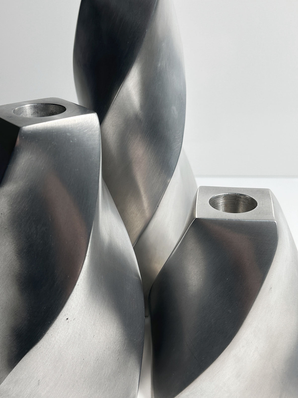 Close-up of three abstract, twisted swirl designs from the "Set of 3 Aluminium Twisted Candle Holders" by Dodo Vintage, featuring smooth surfaces with a brushed metallic finish. Each piece resembles a sculpture with a small circular hole at the top, set against a plain white background.