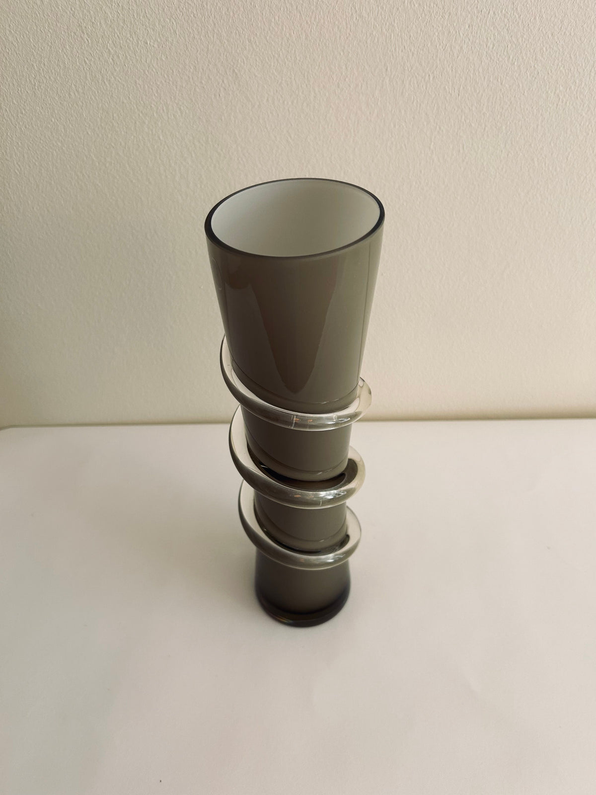 A one-of-a-kind, contemporary vase from Flock Studio, featuring a gray hourglass shape with three horizontal rings of smokey glass spiraling around its center, elegantly displayed against a simple light background.